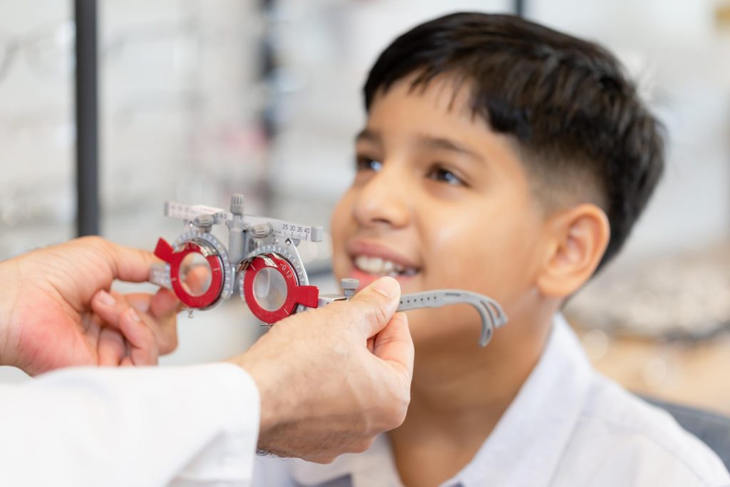 Child seated for an eye exam as a clinician adjusts a red-and-gray trial frame on his face.