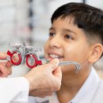 Child seated for an eye exam as a clinician adjusts a red-and-gray trial frame on his face.