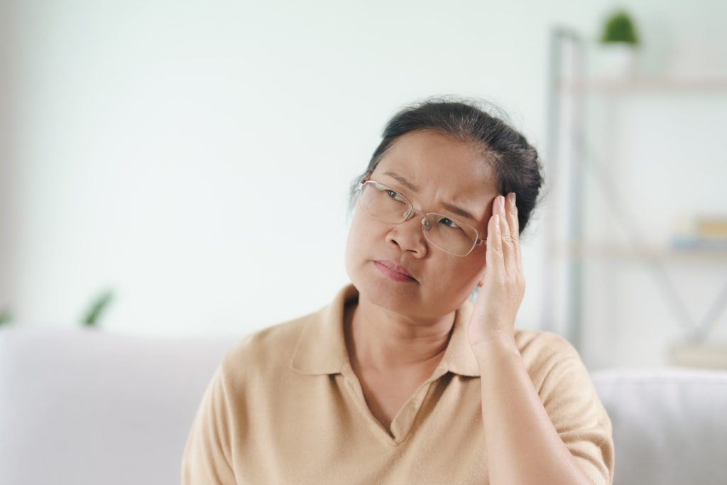 Woman wearing glasses sits on a sofa, touching her temple with a puzzled expression, indoors.